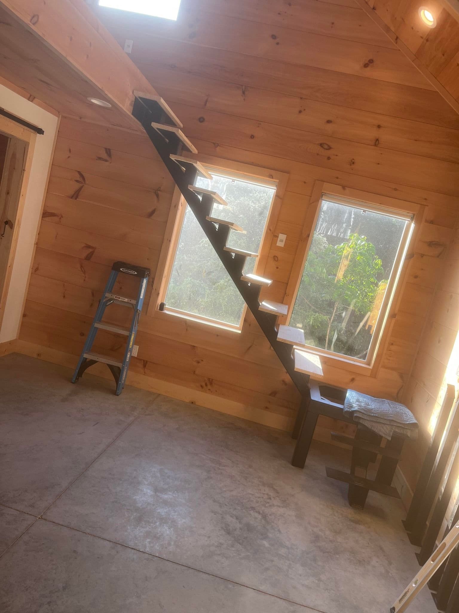 Interior of a wooden tiny house featuring a staircase, large windows, and a ladder.